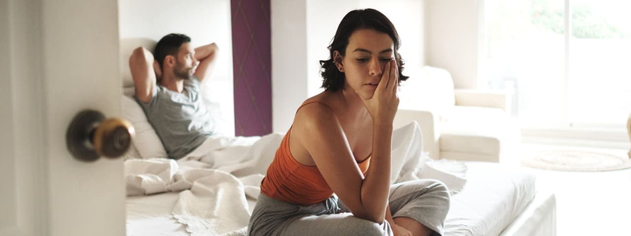 A woman sitting at the end of a bed looking frustrated