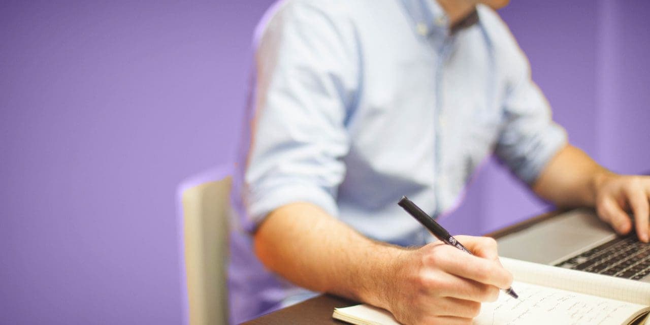 closeup of man infront of a laptop while typing and taking some notes in a notebook